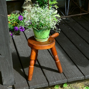 May include: A small, three-legged wooden stool with a round seat and a handle on one side. A light green, leaf-shaped ceramic planter sits on the stool, containing a bushy plant with small white flowers and green foliage.
