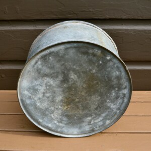 May include: A vintage galvanised metal bucket with a worn, weathered surface. The bucket is round and has a slightly dented bottom.
