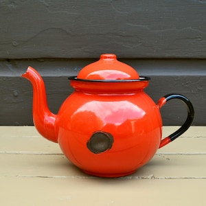 May include: A bright orange enamel teapot with a black rim and handle. The teapot has a small black circle on the front.