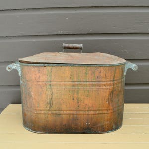 May include: A large, antique copper tub with a matching lid. The tub has two handles on the sides and a wooden handle on the lid. The copper has a weathered, aged appearance, with visible patina and discoloration.