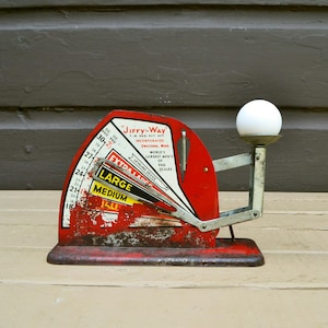 May include: A vintage red metal Jiffy-Way egg scale with a white egg resting on the weighing platform. The device features a curved dial with black text indicating weight categories including Large and Medium, and a metal arm mechanism.