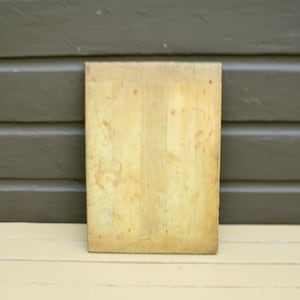 May include: A light brown wooden cutting board with a smooth surface. The board has a worn, vintage look.