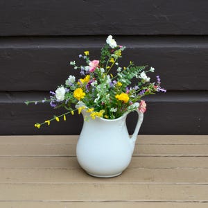 May include: A white ceramic pitcher filled with a vibrant arrangement of wildflowers, including yellow, pink, white, and purple blossoms mixed with green fern-like foliage. The pitcher features a curved handle and a smooth, rounded body design.