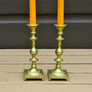 May include: Two matching brass candlesticks with square bases and turned stems, each holding a tall, solid orange taper candle. The metal has a polished finish with visible decorative rings along the vertical shafts of the candle holders.