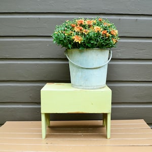 May include: A galvanized metal bucket with a handle holds a vibrant arrangement of orange and yellow chrysanthemums. The bucket sits atop a small, light green wooden step stool. The background is a brown wooden wall.