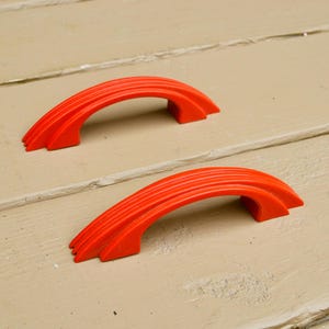 May include: Two orange plastic cabinet door pulls shaped like arches. The pulls are layered to create a three-dimensional effect.