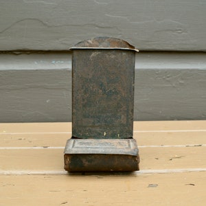 May include: A vintage metal matchbox holder with a worn, dark brown finish. The holder has a rectangular shape with a slightly curved top and a hinged lid.