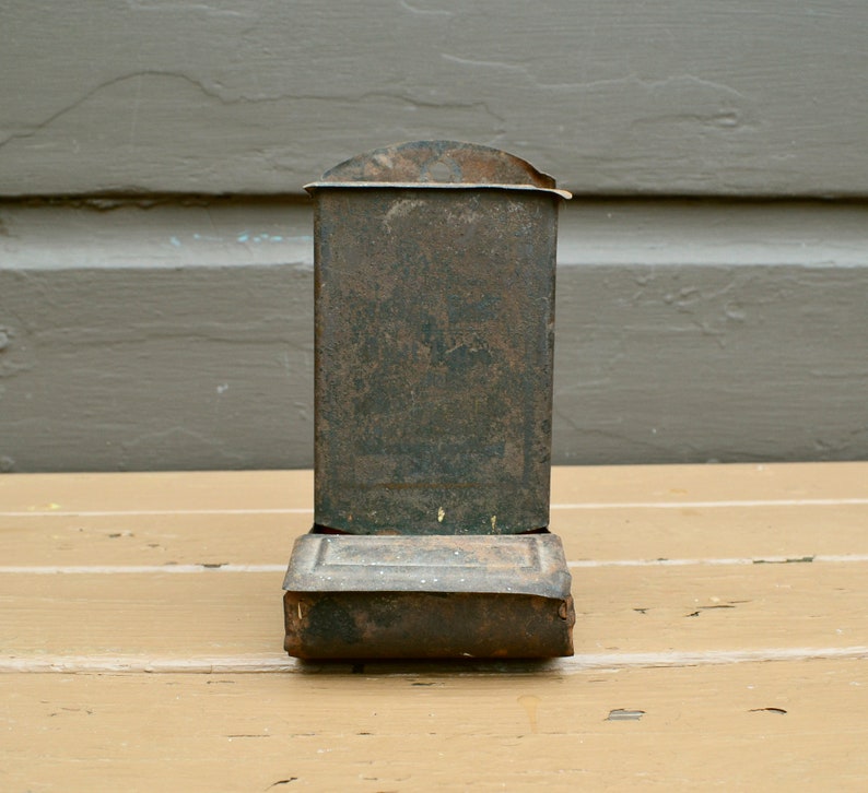 May include: A vintage metal matchbox holder with a worn, dark brown finish. The holder has a rectangular shape with a slightly curved top and a hinged lid.
