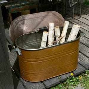 May include: A weathered, oval-shaped metal tub with a copper finish, filled with birch logs. The tub has black handles and sits on a wooden deck. A matching lid rests behind it. The scene evokes a rustic, outdoor theme.