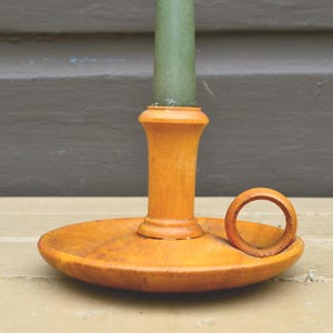 May include: A wooden candle holder with a round base and a central column to hold a green candle. The holder has a warm, light brown color and a small handle on the side. The candle holder is a decorative item.