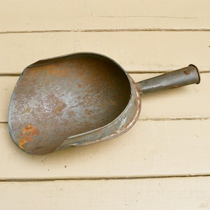 May include: An antique metal scoop with a rounded bowl and a long handle. The scoop has a weathered, rusty appearance with shades of gray and brown, suggesting age and use. The handle is cylindrical and attached to the bowl.