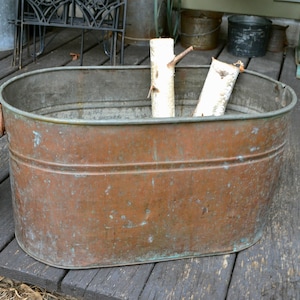May include: A weathered, copper-toned metal tub with an oval shape. Two pieces of birch wood are inside the tub. The tub has a riveted construction and is sitting on a wooden deck.