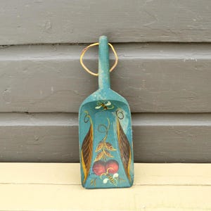 May include: A teal-colored, wooden scoop with a long handle and a gold-colored loop for hanging. The scoop is decorated with painted leaves, berries, and a bee. The scoop is resting on a light-colored surface against a gray background.
