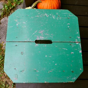 K&ouml;nnte beinhalten: Ein gr&uuml;ner, klappbarer Holzhocker mit einer klappbaren Oberseite und einem ausgeschnittenen Griff. Der Hocker hat ein abgenutztes, verwittertes Aussehen.
