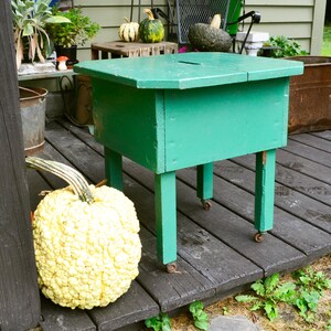 K&ouml;nnte beinhalten: Ein gr&uuml;ner Holztisch mit einer Klappplatte und vier Beinen. Der Tisch hat R&auml;der an der Unterseite der Beine. Ein wei&szlig;er, unebener K&uuml;rbis liegt auf dem Boden neben dem Tisch.