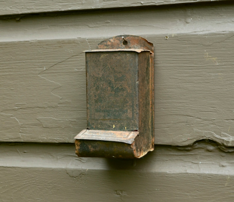 May include: A rusty metal matchbox holder attached to a wooden wall. The holder has a hinged lid and a small compartment for matches.
