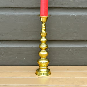 May include: A gold metal candle holder with a decorative, ornate design. The holder has a tall, slender base with multiple tiers of rounded knobs. The candle holder is empty.