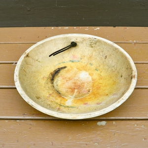 May include: A large, round, wooden bowl with a worn, yellowed finish. The bowl has a dark brown stain on the inside and a small, black metal loop attached to the rim.