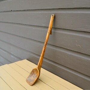 May include: A long, hand-carved wooden spoon with a deep bowl and a slender handle. The spoon is made from a light-colored wood with visible grain and a natural finish. The spoon is leaning against a gray wall and resting on a yellow table.