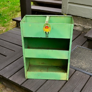 May include: A light green metal storage rack with three open shelves and a central divider. The top front panel features a small, colorful floral decal with red and yellow flowers. The structure is rectangular with a weathered finish.