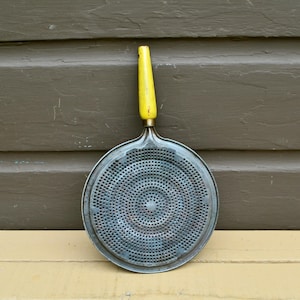 May include: A round, metallic food mill strainer featuring a perforated surface with a dense pattern of small holes. The kitchen tool includes a single, elongated handle finished in a bright yellow color with visible signs of wear and paint chipping.