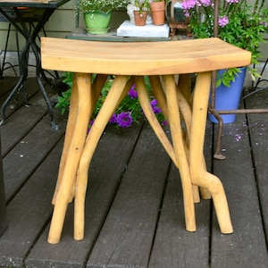 May include: A light brown wooden stool featuring a smooth, rectangular, slightly curved seat supported by four natural, branch-like legs. The stool is constructed from light-toned wood with a visible grain, showcasing a rustic, organic aesthetic.