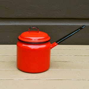 May include: A vibrant red enamelware pot with a lid and a long, black handle. The pot has a black rim and handle accents. This vintage kitchenware item is likely used for cooking or serving.