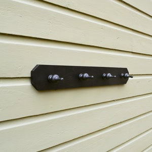 May include: A dark stained wooden coat rack with four metal hooks. The rack is mounted on a light yellow-green horizontal siding. The hooks are evenly spaced along the length of the rack.