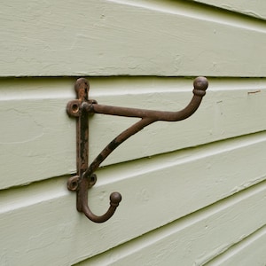 May include: A rustic, brown metal wall hook with two curved arms and a vertical mounting plate. The hook is mounted on a light green wooden wall, showcasing its aged, weathered appearance. The design is simple and functional.