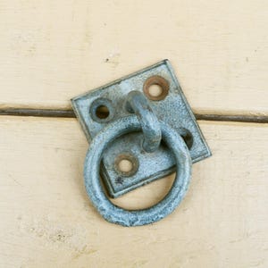 May include: A weathered, square metal ring plate with a circular ring attached. The plate has four holes and a distressed, blue-gray finish. The ring is also metal and shows signs of age and wear.