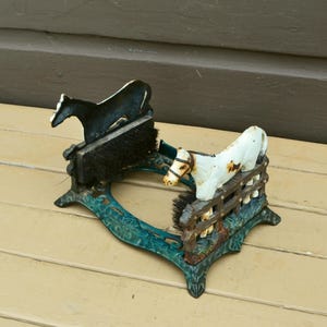 May include: An antique cast iron boot scraper with a horse and fence design. The boot scraper is painted in shades of black, white, and teal. The boot scraper has two brushes for cleaning boots.