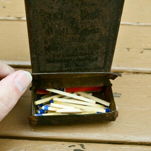 May include: A vintage metal matchbox with a worn, rusty exterior. The box is open, revealing a full stack of wooden matches with blue tips.