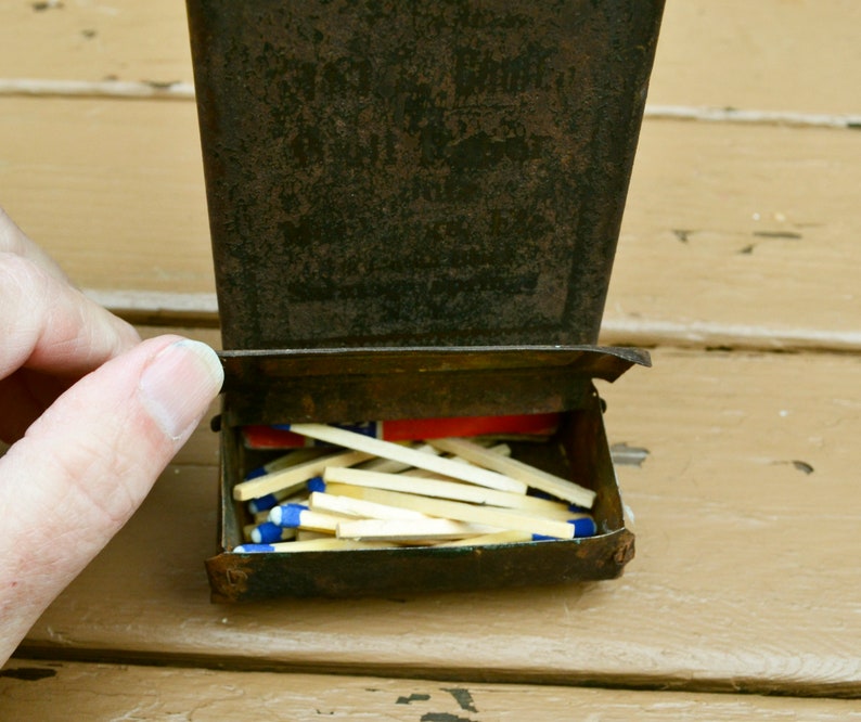 May include: A vintage metal matchbox with a worn, rusty exterior. The box is open, revealing a full stack of wooden matches with blue tips.