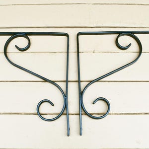May include: Two black wrought iron wall brackets with a decorative scroll design. Each bracket has a horizontal bar and a vertical support with a curved flourish at the top and bottom. The brackets are mounted on a light-colored, horizontal wooden surface.