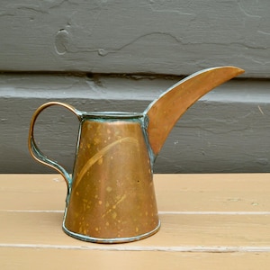 May include: A copper watering can with a long spout and a handle. The can has a patina finish and is slightly tarnished.