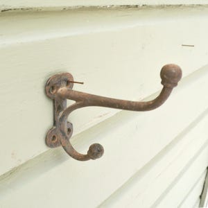 May include: A rustic, antique-style coat hook made of weathered metal. The hook has two curved arms with ball-shaped ends and a decorative mounting plate. The metal has a brown, aged appearance, suggesting it's an older piece.
