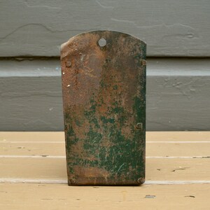 May include: A vintage metal wall-mounted storage unit with a green and brown patina. The unit has a single hole at the top and two small metal tabs on the sides.