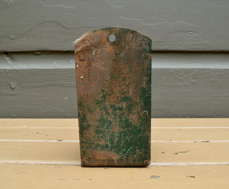 May include: A vintage metal wall-mounted storage unit with a green and brown patina. The unit has a single hole at the top and two small metal tabs on the sides.