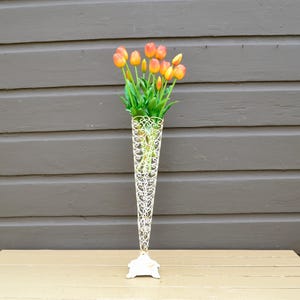 May include: A tall, cream-colored metal vase with an openwork design, holding a bouquet of orange and yellow tulips with green stems and leaves. The vase has a flared top and a decorative base, set against a wooden surface and a gray background.