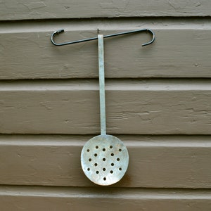 May include: A silver metal slotted spoon with a black metal handle hanging on a brown wooden wall.