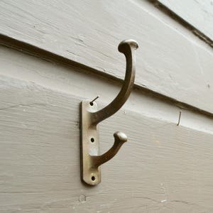 May include: A vintage brass double hook mounted on a weathered, gray wooden surface. The hook has a curved design with two rounded ends, perfect for hanging coats or other items. The hook is attached with screws.