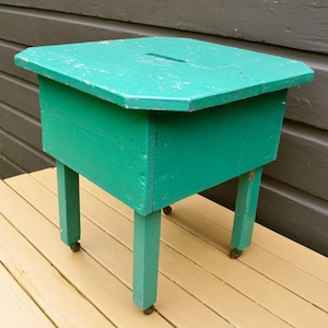 K&ouml;nnte beinhalten: Ein t&uuml;rkisfarbener Holzhocker mit quadratischer Oberseite und vier Beinen. Der Hocker hat ein kleines Loch oben und steht auf einem Holzdeck.