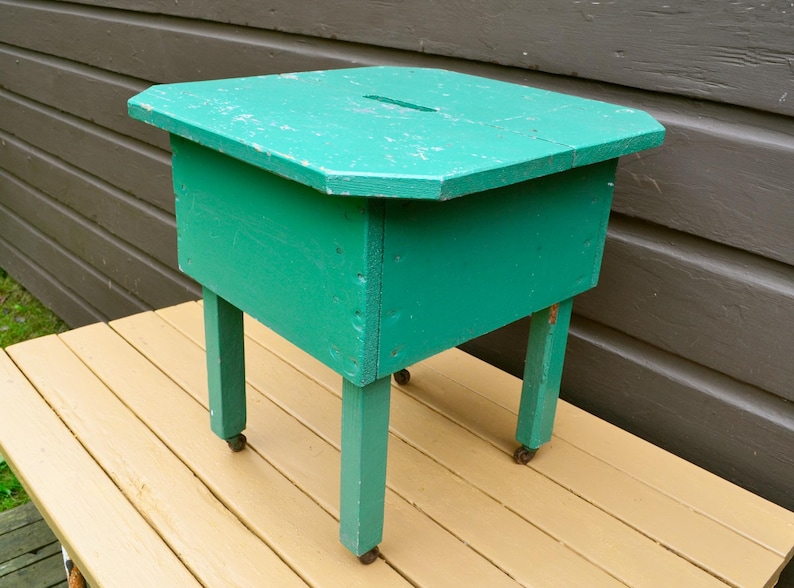 K&ouml;nnte beinhalten: Ein t&uuml;rkisfarbener Holzhocker mit quadratischer Oberseite und vier Beinen. Der Hocker hat ein kleines Loch oben und steht auf einem Holzdeck.