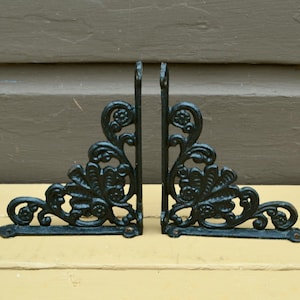 May include: Two black metal shelf brackets with a decorative floral design. The brackets are angled and have a vintage look.