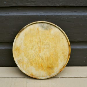 May include: A circular wooden serving tray with a light tan finish, featuring a raised rim and a surface marked by numerous visible scratches and natural wood grain patterns, showing signs of wear and use across the entire flat surface.