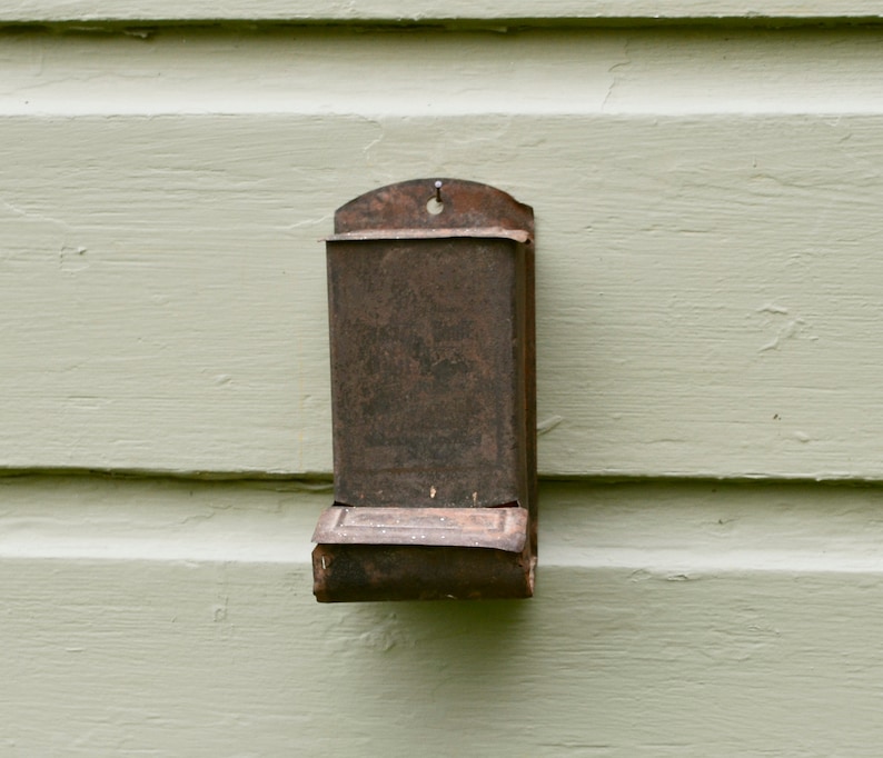 May include: A rusty metal matchbox holder attached to a light green wooden wall.