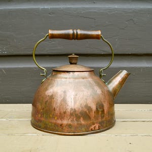 May include: A round copper tea kettle with a dark brown wooden handle and a matching copper lid. The kettle features a curved spout and a brass-toned metal frame connecting the handle to the main body, showing a weathered patina finish.