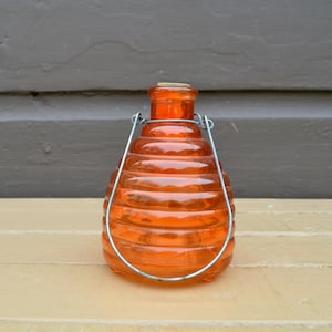May include: An orange glass jar with a beehive design and a metal handle. The jar is made of transparent orange glass and has a series of horizontal ridges. The metal handle arches over the top of the jar.