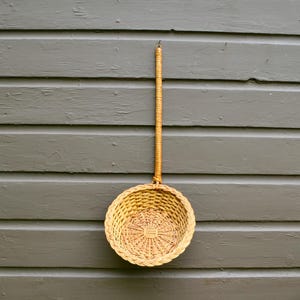 May include: A light brown woven wicker basket with a long, slender handle. The basket features a circular, deep bowl with a braided rim and a flat, woven base. The handle is wrapped in thin wicker strips and extends vertically from the rim.