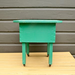 K&ouml;nnte beinhalten: Ein kleiner gr&uuml;ner Holztisch mit quadratischer Tischplatte und vier Beinen. Der Tisch hat R&auml;der an der Unterseite jedes Beines.
